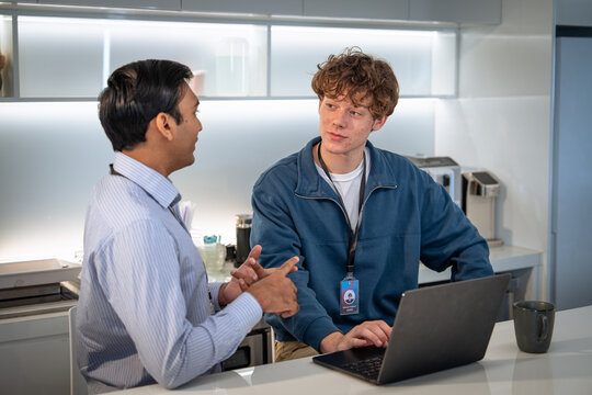 Two men engage in a conversation in a modern workspace, one seated with a laptop and the other standing, both appearing focused and collaborative.