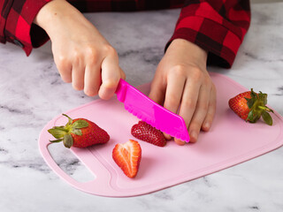 Child cutting strawberries with plastic knife on cutting board, close-up
