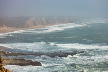 Waves roll toward the long stretch of sandy beach and steep cliffs along California Pacific Coast. A hazy marine layer softens the distant view along scenic Highway 1