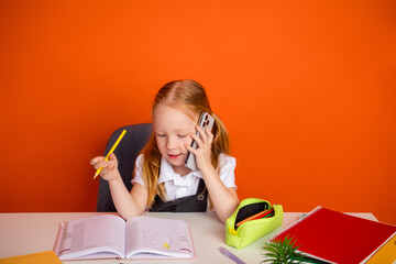 Young adorable schoolgirl on phone while studying with books and pencil against orange background,...