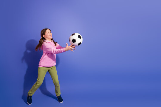 Cheerful schoolgirl in casual fashion catching a soccer ball on a purple background, symbolizing active childhood joy
