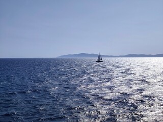Photo of the Aegean Sea under a brilliant summer sky, golden sunlight dancing on the waves, while in the distance a solitary sailboat drifts peacefully across the shimmering blue horizon.