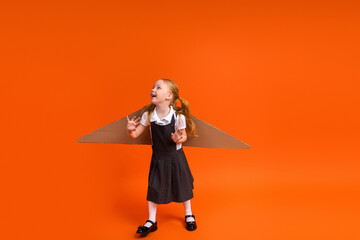 Delighted young girl with cardboard wings in uniform on vivid orange background celebrates learning...
