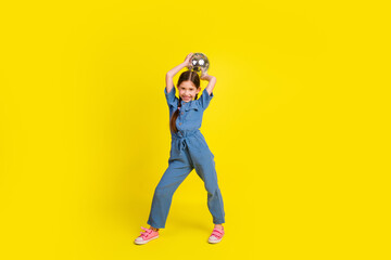 Smiling little girl holding a disco ball against a vivid yellow background, joyfully posing in...