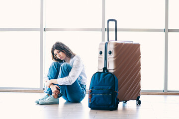 Young woman sitting on the floor with luggage at an airport terminal, waiting for departure and air...