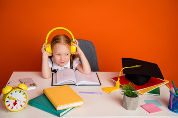 Smiling schoolgirl with headphones studying at desk with school supplies against vibrant orange...