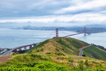 Panoramic view of the Golden Gate Bridge spanning San Francisco Bay with cityscape, lush hills, and boats, captured from a high vantage point on a cloudy spring day