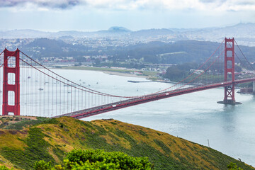 Panoramic view of the Golden Gate Bridge spanning San Francisco Bay with cityscape, lush hills, and boats, captured from a high vantage point on a cloudy spring day