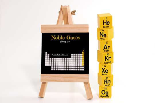 Six yellow cubes, each labeled with the symbols and atomic numbers of noble gases, arranged beside a chalkboard displaying the Periodic Table of Elements, with Group 18 highlighted in yellow