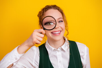 Young woman holding a magnifying glass in front of her eye on a vibrant yellow background...