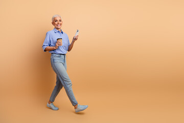 Confident mature woman walking with coffee cup and smartphone on beige background demonstrating...