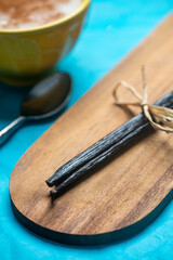 Close up studio still life photo featuring dried vanilla beans on a vintage wooden board and vibrant blue hand made paper background with a bowl of rice pudding sprinkled with cinnamon