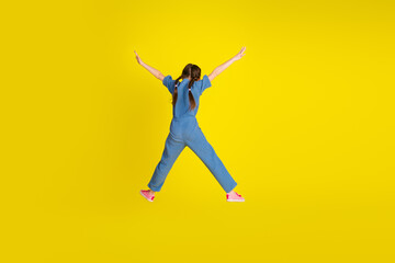 Young girl joyfully jumping in casual blue outfit against vibrant yellow background expressing happiness and energy