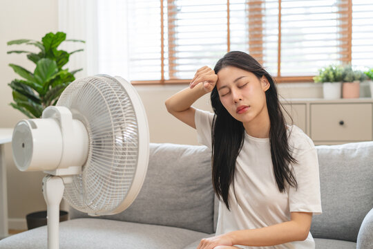 Suffer summer heat wave stroke, hot weather, tired asian young woman sitting on couch in living room without air-conditioner, using electric fan, wave fan to cool for ventilation when high temperature - Powered by Adobe