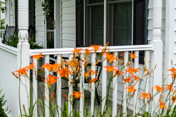 inviting front porch of white colonial with black shutters white railing black rocking chair and bench through out of focus orange flowers
