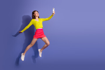 Young woman in vibrant outfit taking photo with phone against purple background conveying fashion...