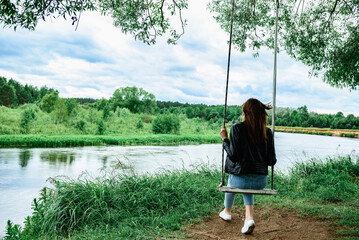 A happy young girl is sitting on a swing in a beautiful tree by the river.