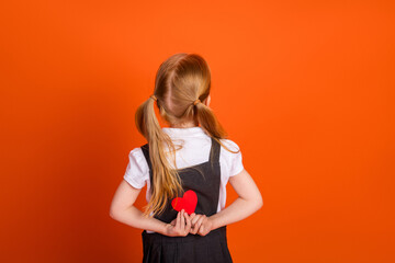Young girl with red hair holding a red paper heart while standing against an orange background in a...