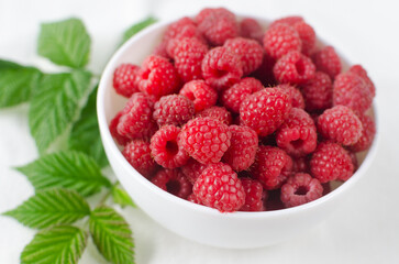 Ripe pink raspberries in a bowl with green leaves on a white background. Concept of summer berry harvest. Horizontal orientation. Selective focus.