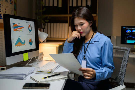 Asian businesswoman working late and reviewing paperwork showing concern about financial charts on computer screen