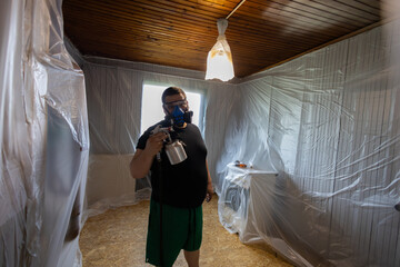 A male painter wearing a protective mask and using a paint sprayer repairs a room covered with...