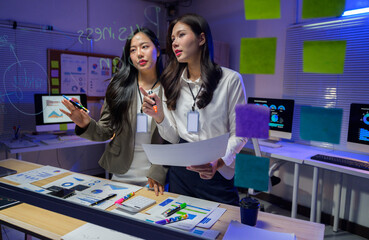 Two businesswomen analyzing graphs and charts on a transparent digital screen using sticky notes in a modern office working late at night