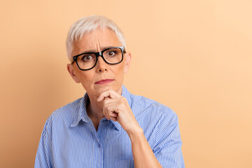 Senior Businesswoman in Thoughtful Gesture Against Beige Background, Wearing Glasses and Striped Blue Shirt