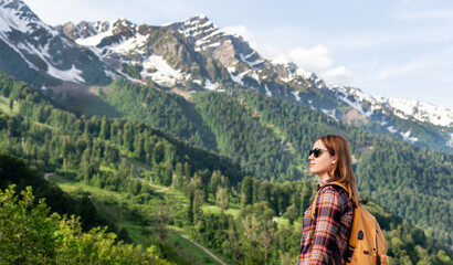 Young woman hiker with sunglasses and a backpack stands in a lush green valley, gazing at majestic...