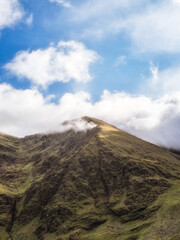 A majestic mountain rises towards a vibrant blue sky dotted with fluffy white clouds. The peak is...