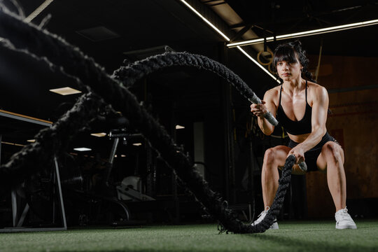 Strong woman using battle ropes for intense gym workout motivation in modern fitness training studio