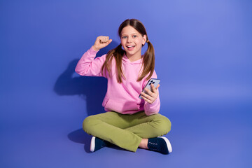 Happy schoolgirl in casual outfit sitting with smartphone showing cheerful celebration on purple...