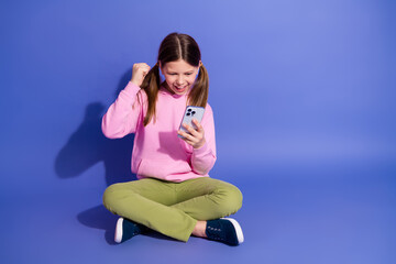 Excited young girl in a pink hoodie celebrates while using a smartphone on a purple background,...
