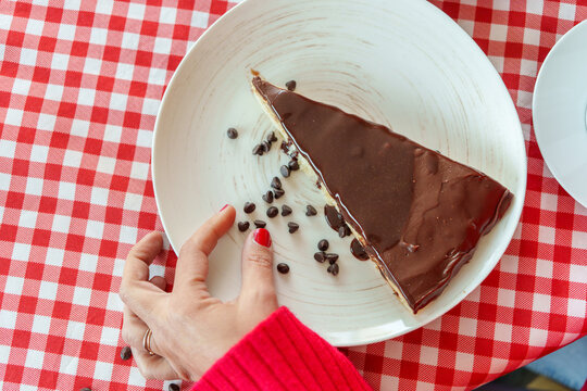 Woman in red sweater reaching for chocolate chips resting on edge of chocolate cake slice, placed on white plate over checkered red white tablecloth