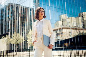 Confident businesswoman in white suit standing outside modern urban building on a sunny day,...