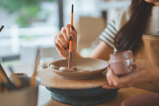 Hands of woman artist painting handmade ceramic product in pottery studio – creative process of decorating sculpture with brush for small business, art workshop, and modern craft design startup