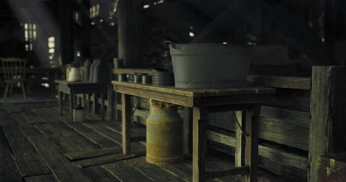 Inside a rustic wooden cabin, kitchen supplies are arranged on tables. The warm light from outside casts shadows, creating a cozy atmosphere at dusk.