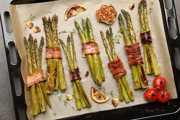 Tasty cooked asparagus with bacon, tomatoes and spices in baking tray on grey table, top view