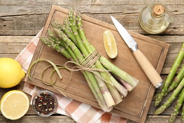 Fresh raw asparagus spears, knife, lemon and oil on wooden table, flat lay