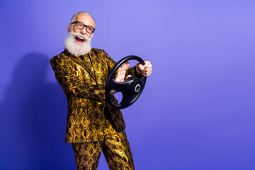Cheerful senior man in glamorous gold attire playfully posing with a steering wheel against a...