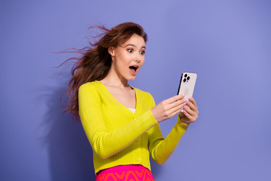 Young woman in vibrant clothing expressing surprise while looking at her smartphone against a purple background