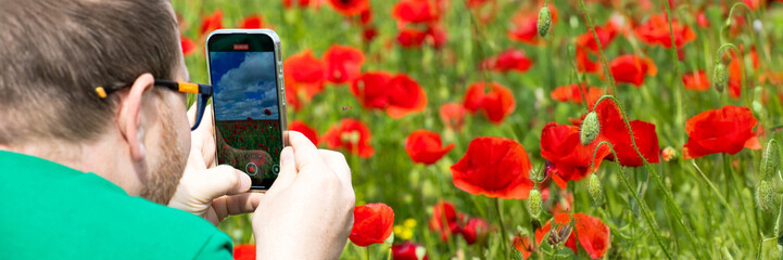 A man with glasses captures blooming poppies on a smartphone during Poppy Day, evoking digital...