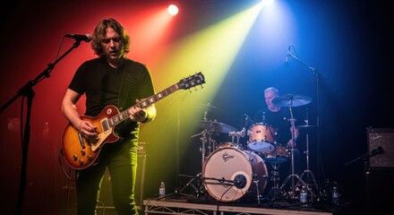Fototapeta premium A male guitarist with long curly hair playing an electric guitar on stage, with a drummer in the background, under vibrant red, yellow, and blue stage lights, capturing a dynamic live performance.