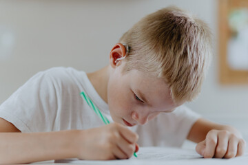 A boy with hearing aids focuses on homework, showcasing natural inclusive education. Authentic...