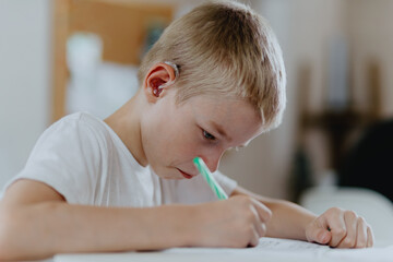 A boy with hearing aids focuses on homework, showcasing natural inclusive education. Authentic...