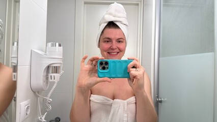 Smiling Eastern European woman captures towel-clad selfie, embodying refreshing hygge ritual,...