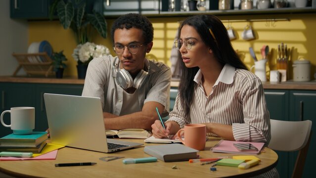 Multiracial woman man studying together at kitchen table high school college students African American guy Latino woman two teenagers use laptop computer online class write homework distant education - Powered by Adobe