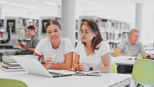  Two positive ladies wearing casual clothes behave loudly while studying in the library and using computer