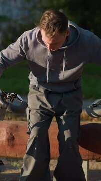 Man outdoors in park walking and sitting on redwood log holding spring stilts, wearing gray hoodie and dark pants, serious expression, surrounded by green grass and trees under bright sunny sky