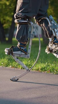 Person using spring stilts on paved path in park, wearing dark gray pants and athletic shoes, captured from low angle focusing on legs and stilts, showcasing dynamic motion and energy