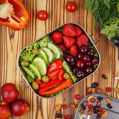 Stainless steel lunch box filled with strawberries, cherries, cucumbers, olives, and bell pepper slices, surrounded by fresh ingredients. A healthy, colorful meal idea on rustic wood background